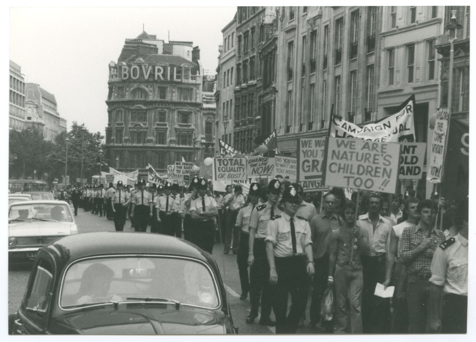 Marș Pride la Londra în 1974, cu manifestanți, polițiști și pancarta „We Are Nature's Children Too”.