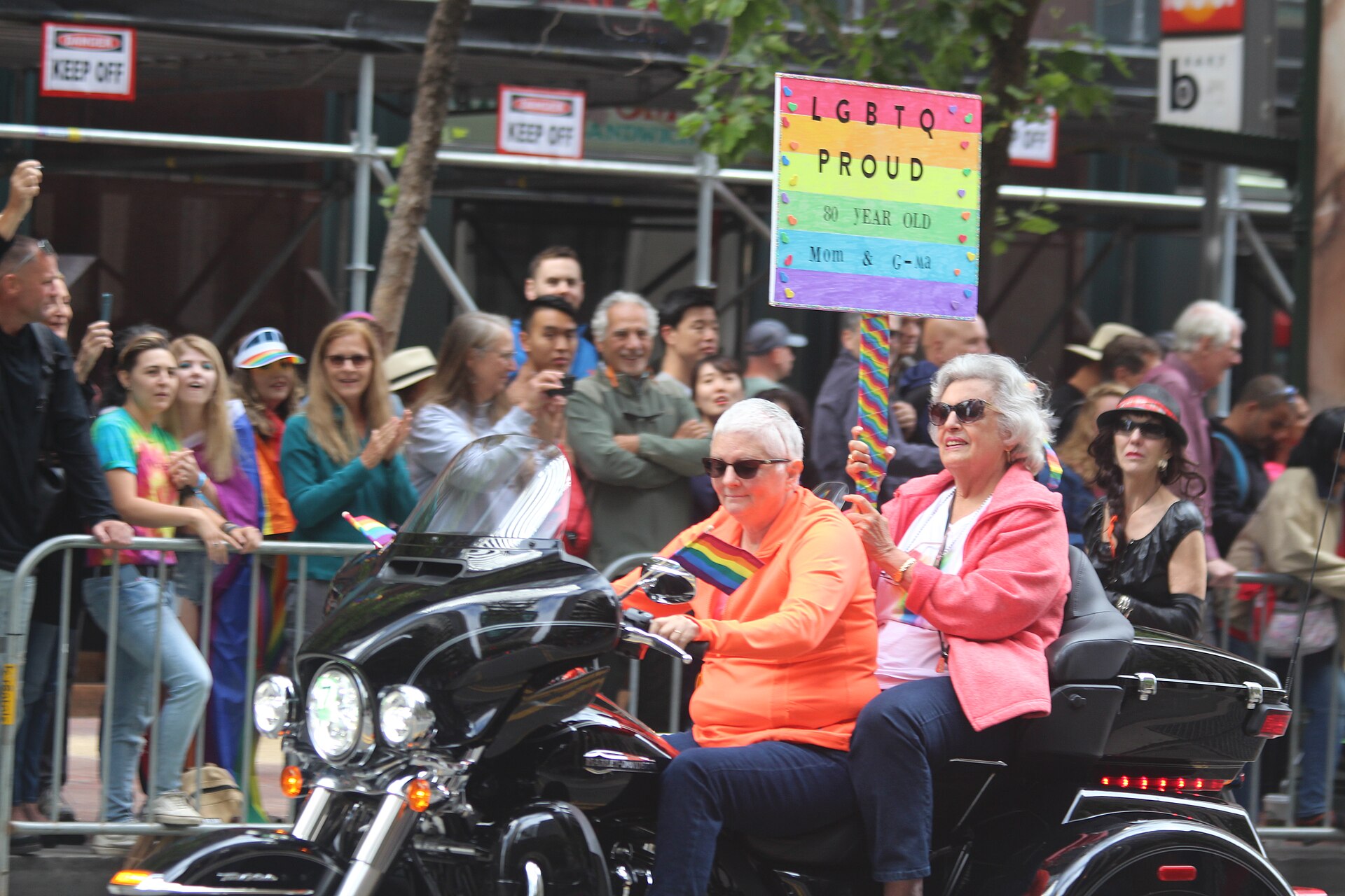 Două participante în vârstă la San Francisco Pride 2019, pe o motocicletă, una ținând o pancartă „LGBTQ Proud”.