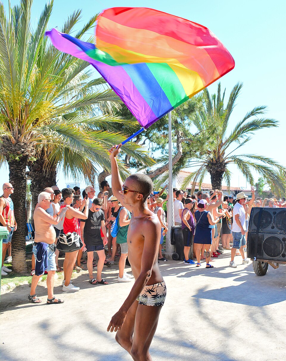 Un steag curcubeu flutură deasupra mulțimii, printre palmieri, la Sitges Pride 2017.